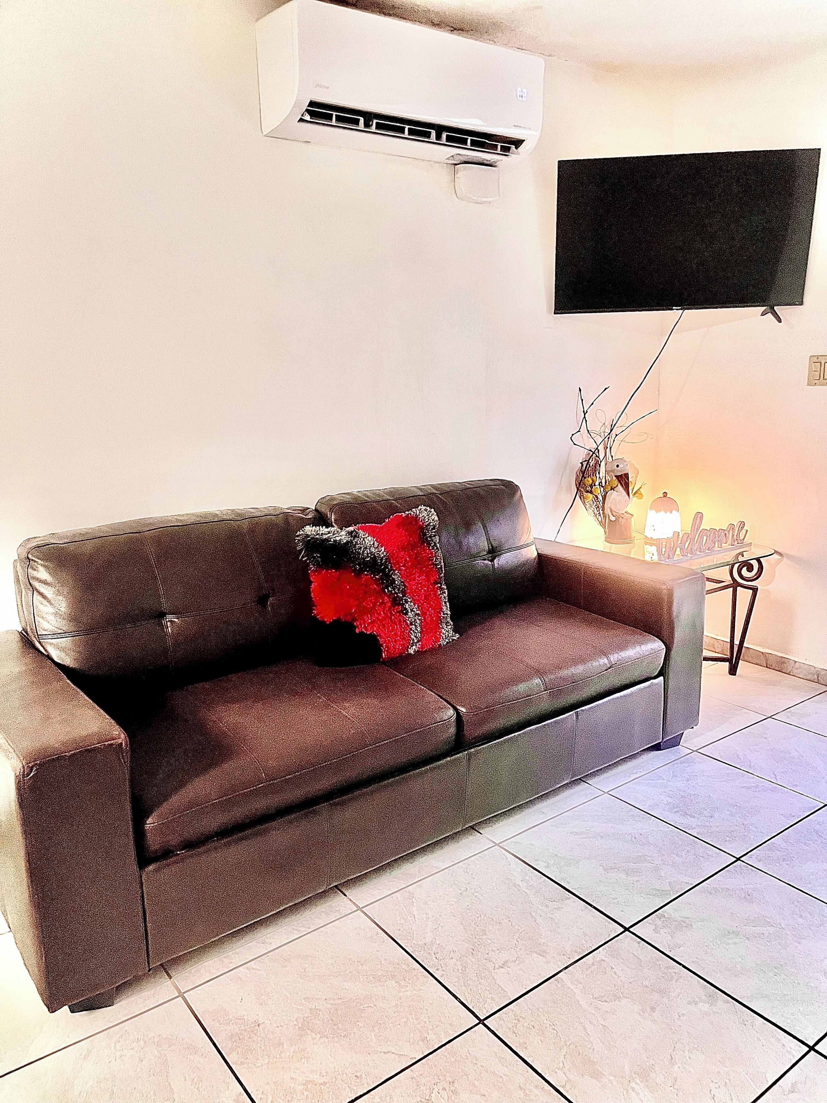Un sofá de cuero marrón con un cojín rojo y negro, un aire acondicionado instalado, un televisor de pantalla plana en la pared y una mesita con una lámpara y un adorno.