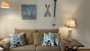 Un salon avec un canapé beige, deux coussins à motifs, un tableau représentant des voiliers, une pagaie et une lampe.