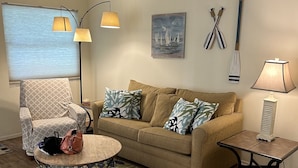 Un salon avec un canapé beige, un fauteuil à motifs, une table basse ronde et une petite table d’appoint avec une lampe.