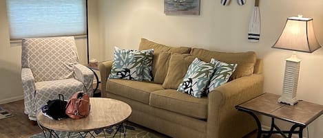 A living room with a beige sofa, patterned armchair, round coffee table, and a small side table with a lamp.