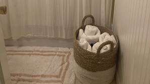 Racó d'una habitació amb una porta blanca, una cortina blanca, una cistella teixida amb tovalloles i una catifa blanca amb dibuix de puntes de fletxa.