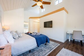 Un dormitorio con cama, escritorio, silla, ventilador de techo y televisor montado en la pared.