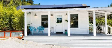 A white building with a covered porch, solar panels on the roof, and two blue chairs outside.