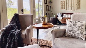Ein gemütliches Wohnzimmer mit Kamin, einem runden Holztisch mit Flasche und Gläsern sowie einer Couch mit einem dekorativen Kissen.