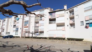 A multi-story residential building with balconies, a parking lot, and a tree in the foreground.