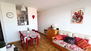 A cozy living room with a wooden floor, a dining area with a red table and chairs, a sofa with patterned cushions, a kitchen area with a clock, and a colorful abstract painting on the wall.