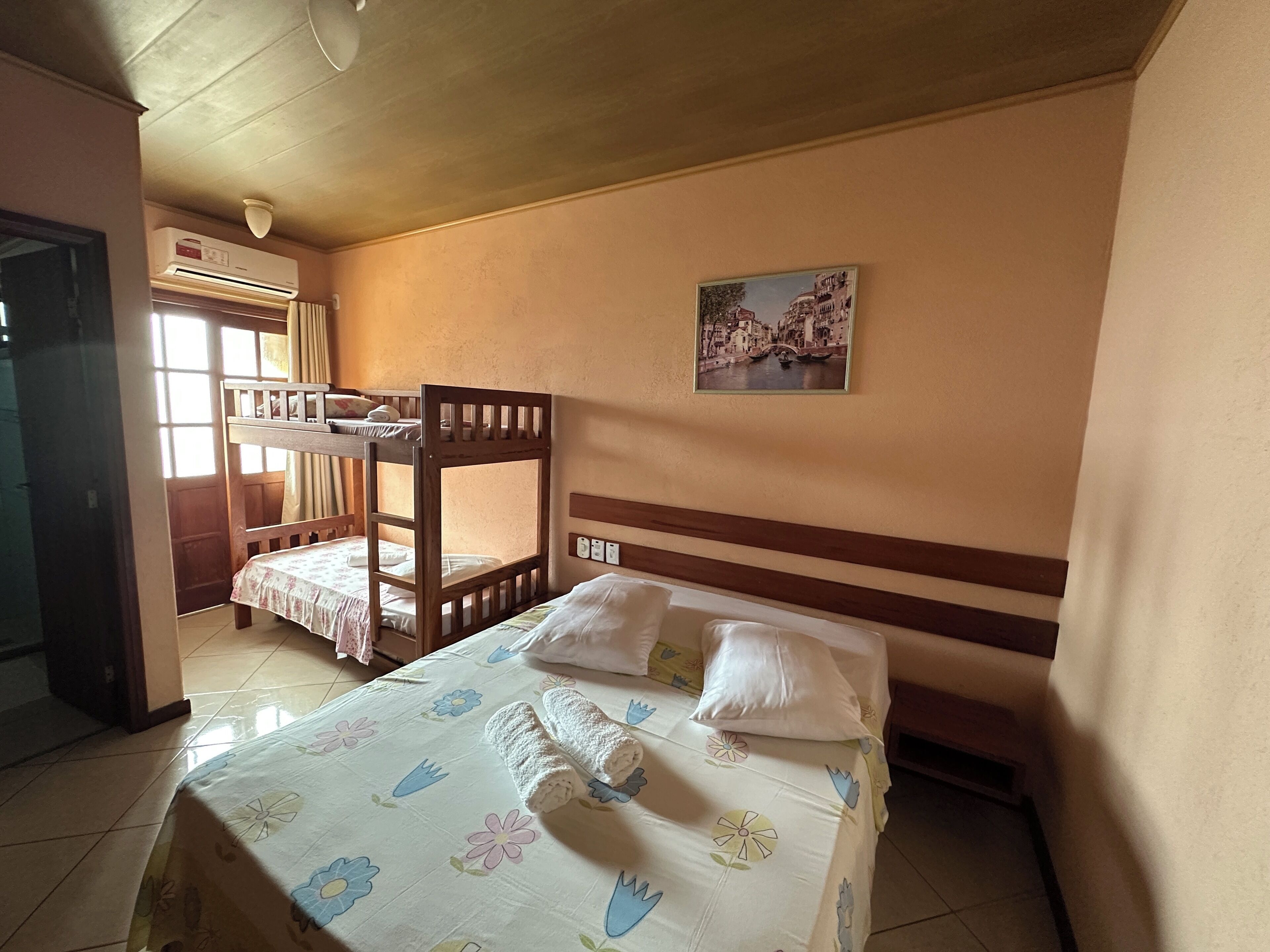 A room with bunk beds, a bed with a floral bedspread, a wall-mounted air conditioner, and a picture of a cityscape.