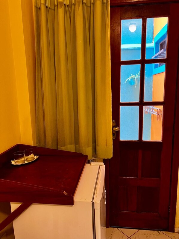 A room with a wooden door, a window with a view of a building, a small table with a tray, and a curtain. - Pousada Maravista (Anchieta)