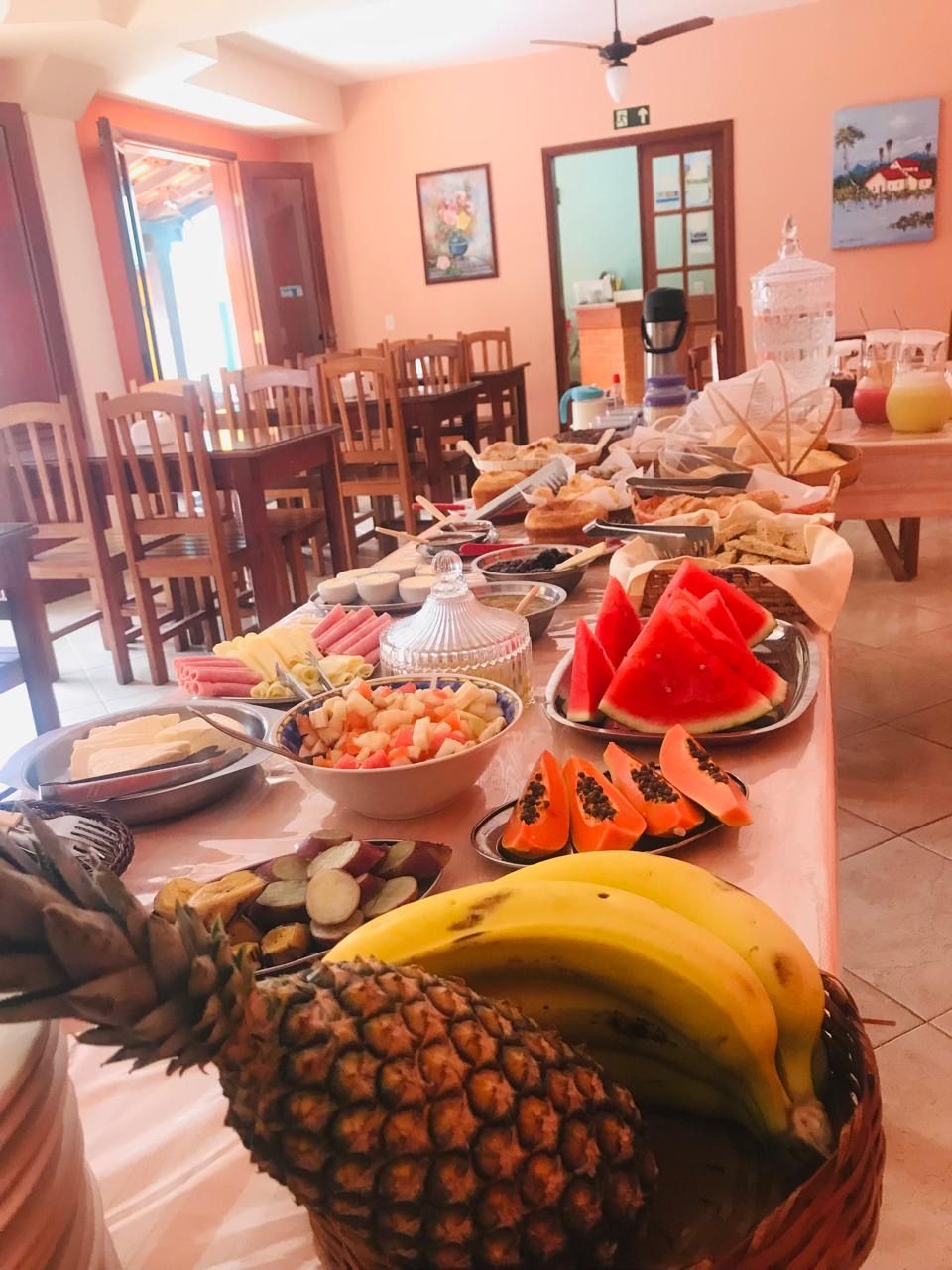 Una mesa de buffet con diversas frutas, como piña, plátanos, sandía y papaya, en una sala con sillas de madera y un ventilador de techo.