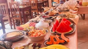 Una mesa de buffet con diversas frutas, como piña, plátanos, sandía y papaya, en una sala con sillas de madera y un ventilador de techo.