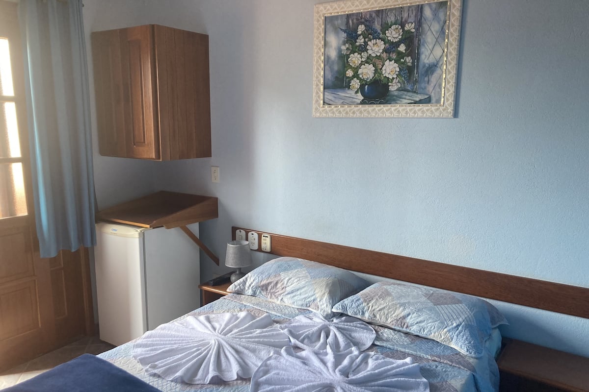 A bedroom with a bed, a nightstand, a lamp, a painting, a wooden cabinet, and a small refrigerator.