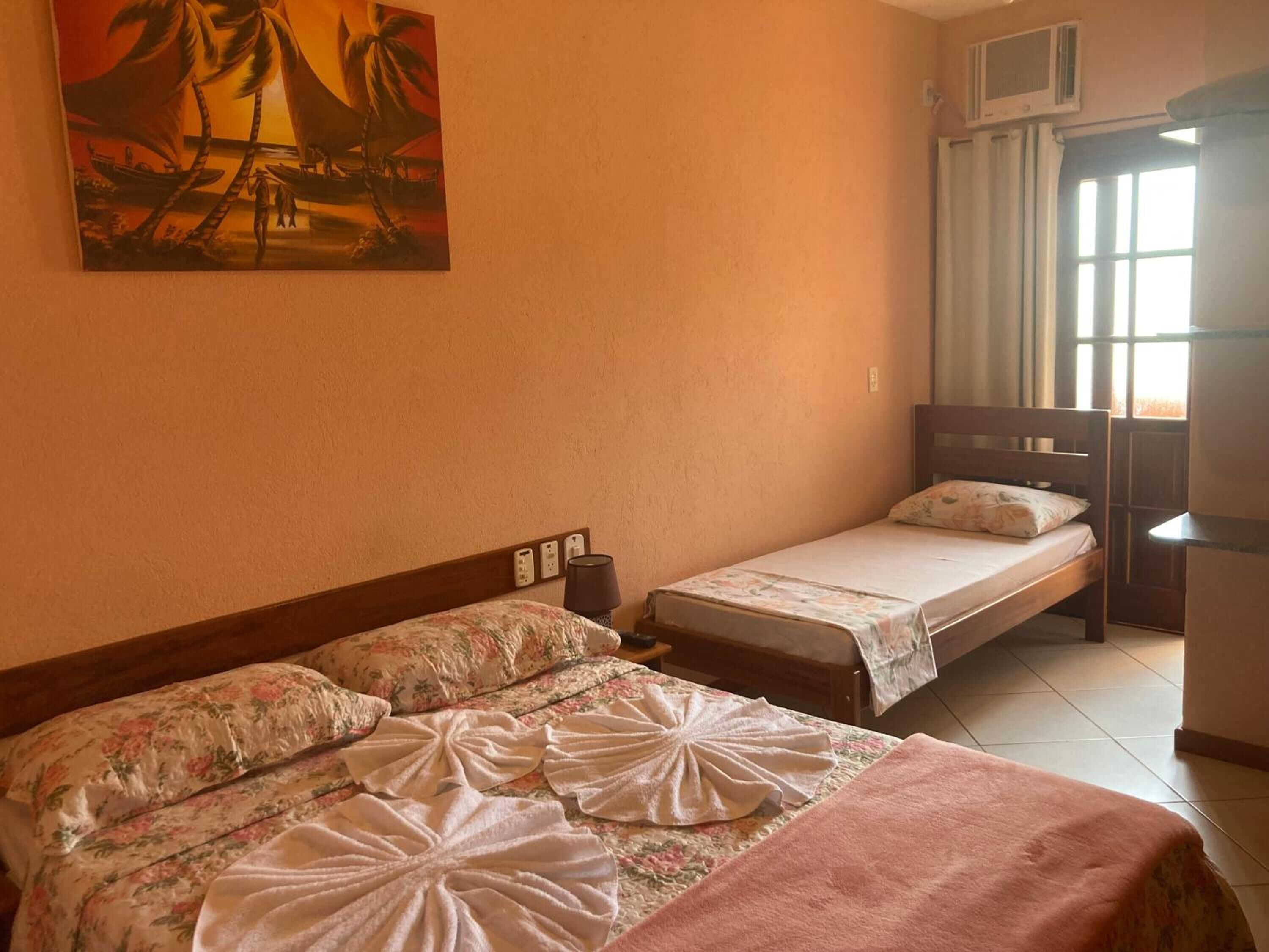 A hotel room with two beds, a painting of palm trees, a window with curtains, and an air conditioning unit.