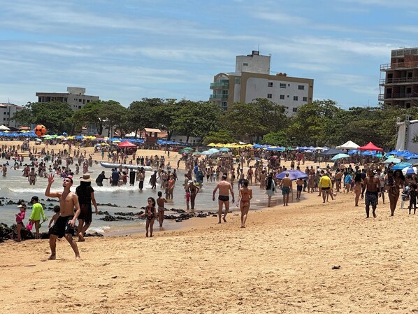 Una playa concurrida donde gente camina y juega en la arena, sombrillas y edificios a la orilla del mar al fondo.