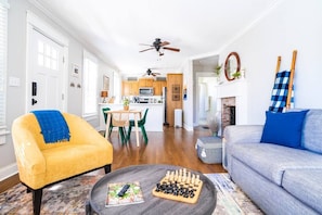 A living room with a yellow armchair, a grey sofa, a coffee table with chess pieces, and a kitchen in the background.