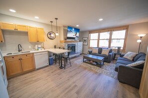 A modern kitchen with wooden cabinets, a dining area with a table and chairs, a fireplace, and a living room with a sofa and a TV.
