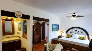 A bedroom with a bed, a nightstand, a lamp, a ceiling fan, a window with a view, and a bathroom with a sink and mirror. - Casa Tortuga @ Selva Romantica (Puerto Vallarta)