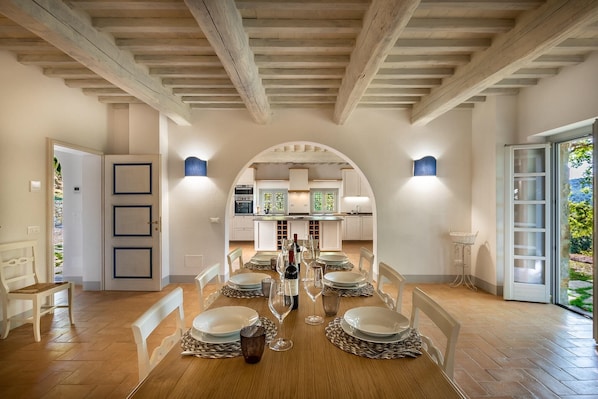 A dining area with a long table set for a meal, a kitchen in the background, and a door leading outside.
