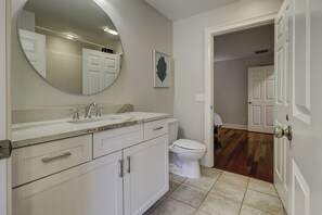 Un baño con tocador blanco, un espejo redondo, inodoro y puerta que da a otra habitación.
