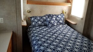 Une petite chambre à coucher, de dimensions réduites, avec un lit agrémenté d’une couvre-lit aux motifs géométriques, une table de chevet et un luminaire fixé au mur.