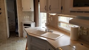 Une petite cuisine fonctionnelle équipée d’un évier à deux bacs, de meubles blancs et d’une fenêtre laissant entrer la lumière naturelle.