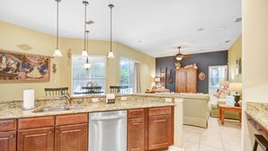 A spacious kitchen with a large island, wooden cabinets, and a dishwasher. There is a dining area with a table and chairs, and a living room with a sofa and a lamp.