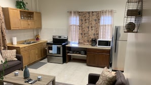 A compact kitchen with a stove, oven, microwave, and sink. There is a wooden cabinet above the sink and a small table with a vase and remote controls.