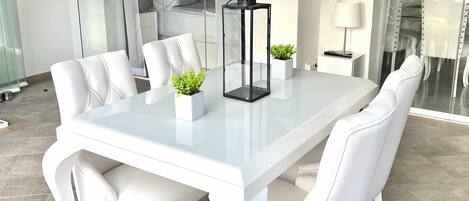 A modern dining area with a glass table, white upholstered chairs, and two potted plants.