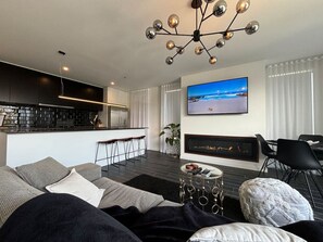 Un salon moderne avec un canapé, une table basse, une cheminée, un téléviseur et, au fond, un coin cuisine.