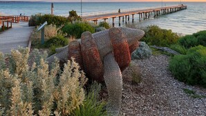 A coastal scene with a wooden pier extending into the sea, surrounded by landscaped gardens and a large, textured sculpture.