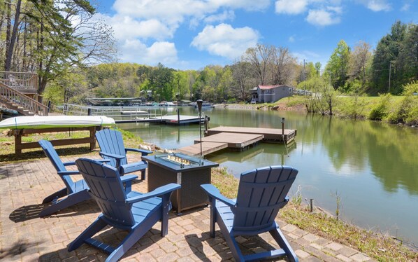 A lakeside patio with Adirondack chairs and a fire pit, overlooking a calm lake with docked boats and a small building in the background.