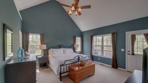 Une chambre à coucher comprenant un lit, des tables de chevet, une commode en bois, un ventilateur de plafond et de grandes fenêtres.