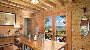 Interior de una cabaña de madera con una mesa de comedor, sillas, una lámpara, un espejo y vistas de montañas a través de una puerta.