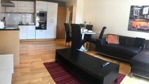 A modern living room with a black sofa, a coffee table, and a dining area with chairs and a table. A kitchen with white cabinets and a microwave is visible in the background. - Apartment-sleeps4-balcony-parking-petsok (Hull)