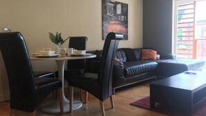 A modern living room with a black leather sofa, a round dining table with chairs, a coffee table, and a large window with a view of a building. - Apartment-sleeps4-balcony-parking-petsok (Hull)