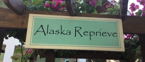 A sign with the text "Alaska Reprieve" on a wooden pergola with green foliage and pink flowers.