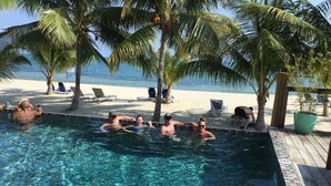 Ein Poolbereich mit klarem Wasser, umgeben von Palmen und einem Sandstrand.