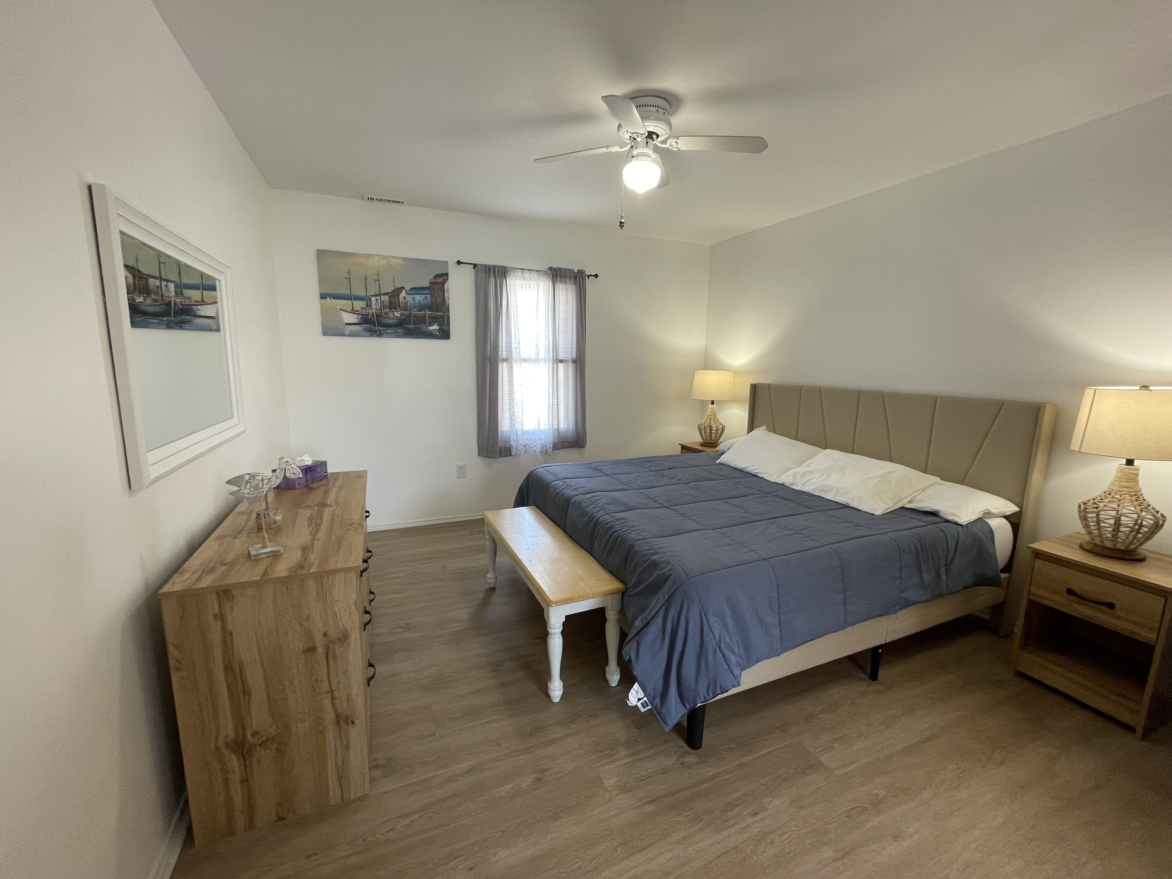 A bedroom with a bed, bedside tables, a desk, a ceiling fan, and artwork on the walls.
