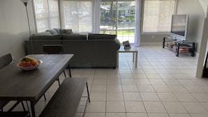 Ein modernes Wohnzimmer mit einer Couch, einem Esstisch mit Obstschale, einem Fernseher auf einem Schrank und großen Fenstern mit Jalousien.