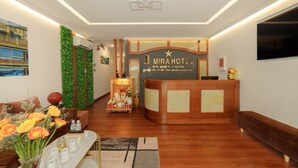 Zona de recepción de un hotel con un mostrador de madera, un televisor montado en la pared y una mesa de recepción.