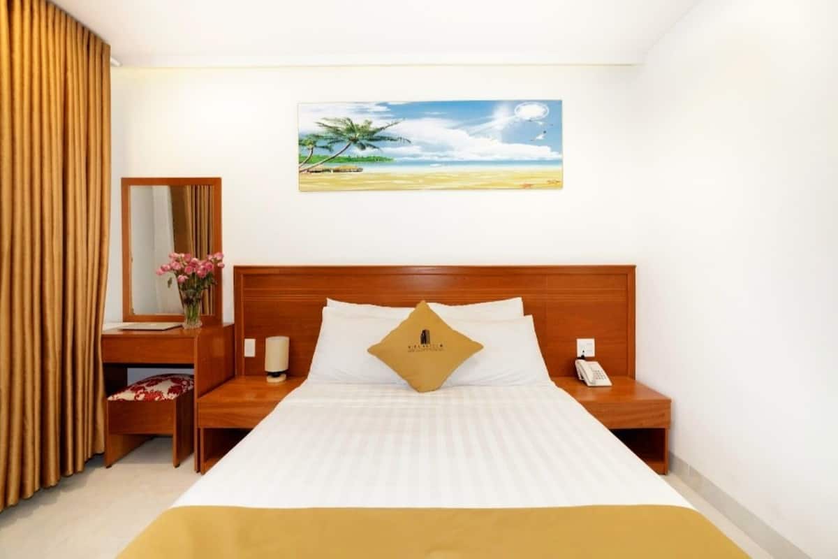 Una habitación de hotel con cama, mesita de noche, escritorio, silla, espejo, un cuadro de una playa y teléfono.