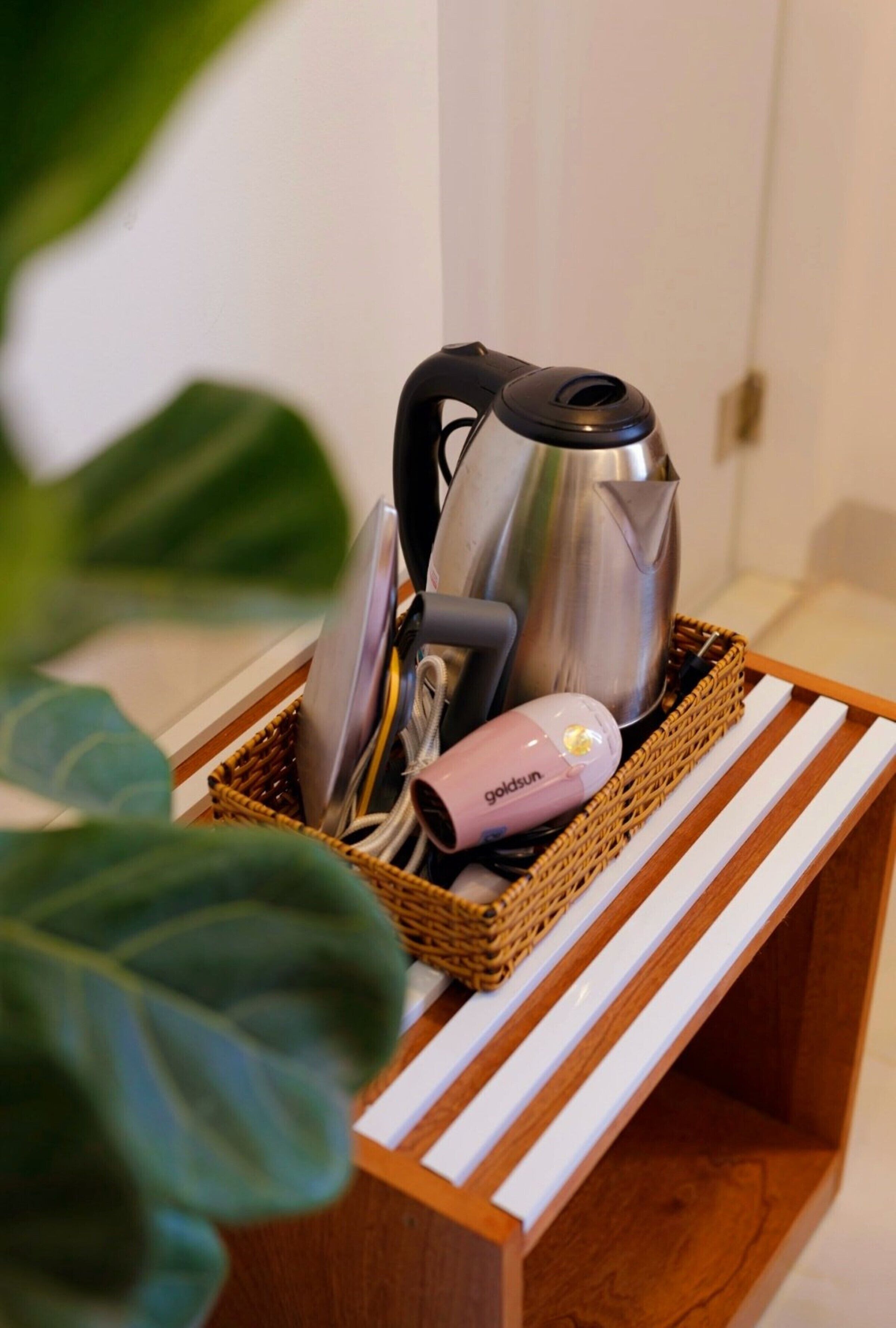 A wicker basket containing a kettle, a pink hairdryer, and a pair of scissors on a wooden shelf.
