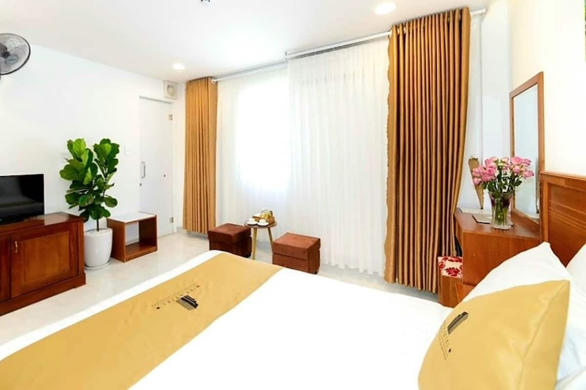 Habitación de hotel con cama, televisor, planta, mesita con un florero y ventana con cortinas.