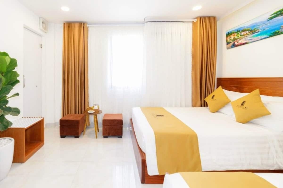 Habitación de hotel con una cama grande, dos mesitas de noche, una mesita pequeña, una planta en maceta y un cuadro en la pared.