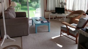 A living room with a sofa, a coffee table, a TV, and a chair. There is a window with curtains and a view of greenery outside. - Farmstay, 2 bedrooms with 2 private baths, private entrance. (Waldoboro)
