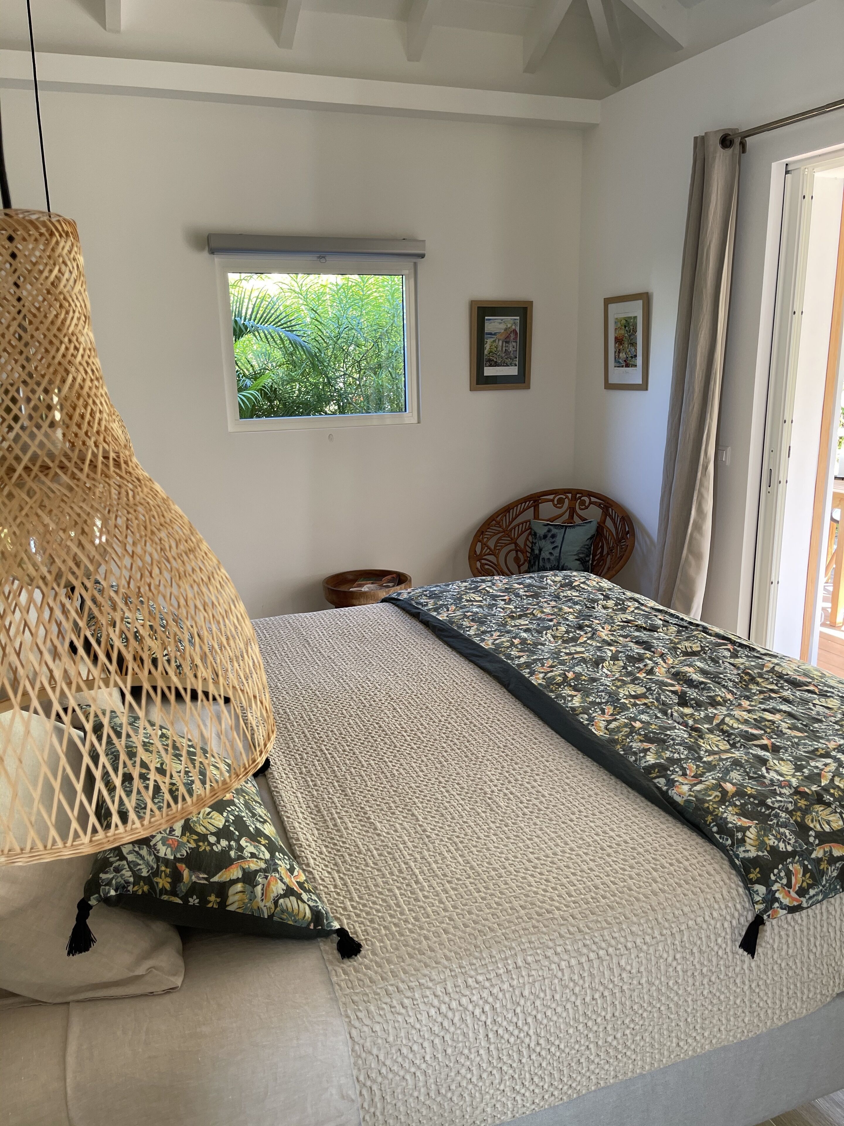 A bedroom with a bed, a woven hanging chair, a window with a view of greenery, and two framed pictures on the wall.
