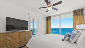Une chambre d’hôtel avec un grand lit, un téléviseur posé sur une commode en bois, un ventilateur de plafond et un balcon offrant une vue sur l’océan.