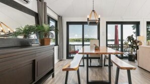 Een moderne eethoek met een houten tafel en banken, grote ramen met uitzicht op het water en een plafondventilator.
