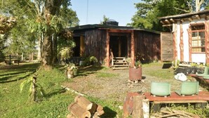 A rustic wooden house with a porch, a small shed, and a garden area with chairs and a table. - The round house in Filandia with waterfall (Filandia)