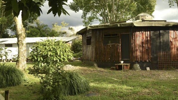 Une cabane en bois rustique, surmontée d’un toit en tôle ondulée, entourée d’une végétation luxuriante et sous un ciel nuageux.
