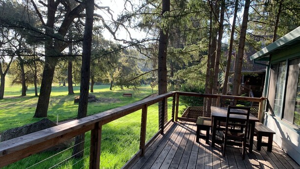 Terraza de madera con mesa y sillas, rodeada por barandilla de madera y con vista a un espacio cubierto de césped y árboles.